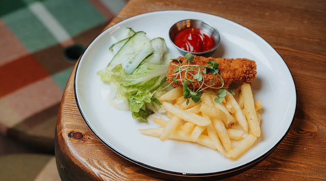 En vit tallrik på ett träbord innehåller panerad kött, pommes frites, sallad och en skål med ketchup.