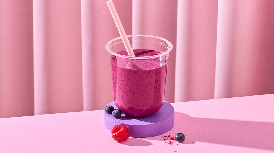 Marjasmoothie kirkkaassa muovimukissa ja pillillä violetilla alustalla. Vieressä mustikoita ja vadelma vaaleanpunaista taustaa vasten.