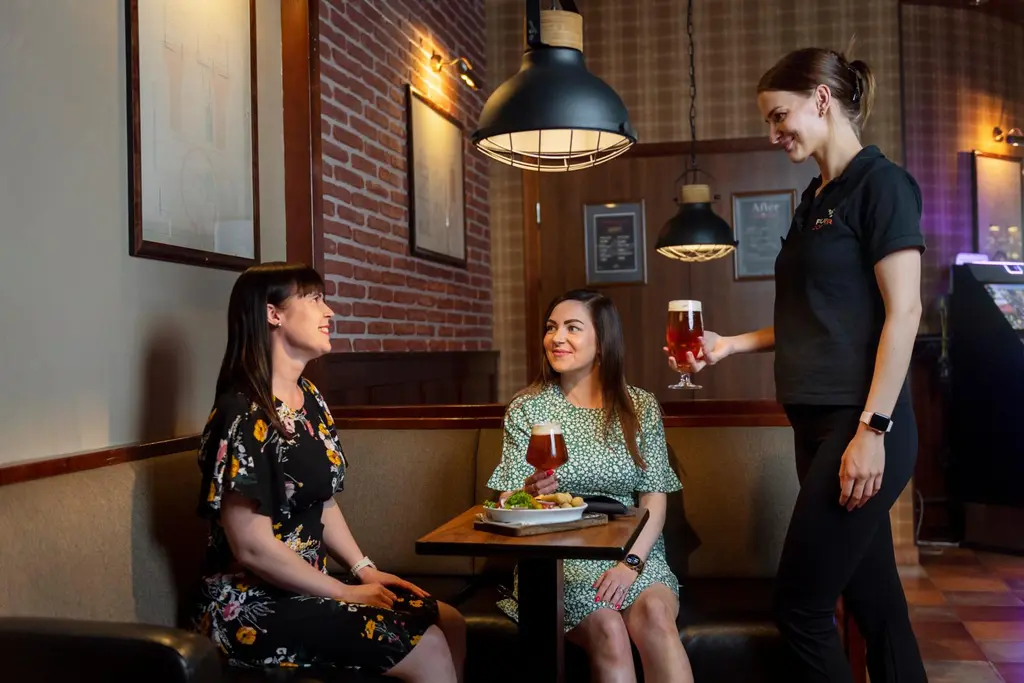 Kaksi nuorta naista istuu ravintolapöydässä juoman ja ruoan ääressä. Tarjoilija tuo lasituopin olutta pöytään.