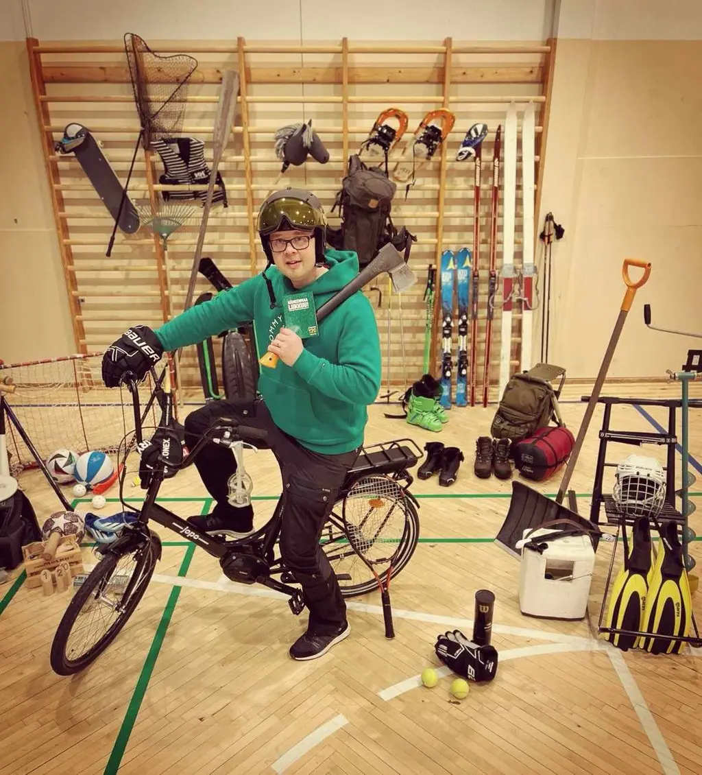 Mies istuu polkupyörän satulassa keskellä liikuntasalia. Ympärillä on kasa erilaisia liikuntavälineitä.