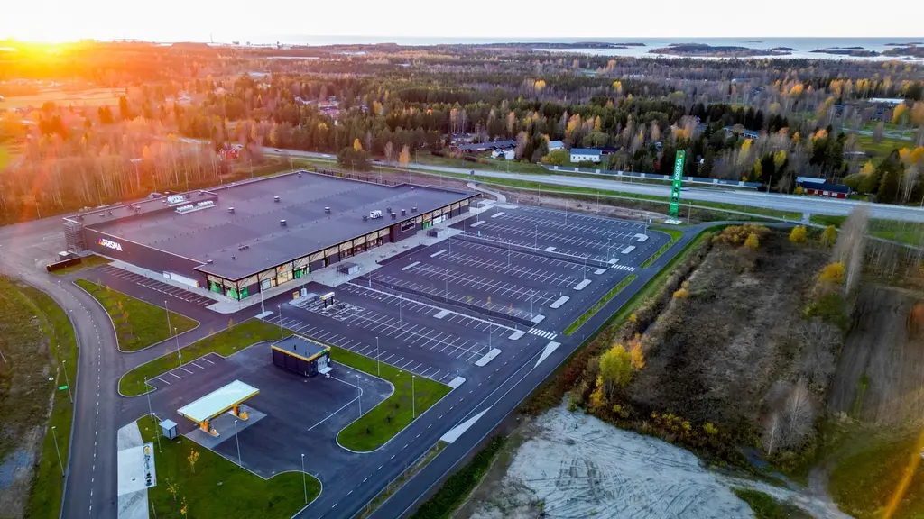 Ilmakuva suuresta, parkkipaikkojen ympäröimästä liiketilasta maaseudun maisemassa. Aurinko paistaa matalalta taustalla.