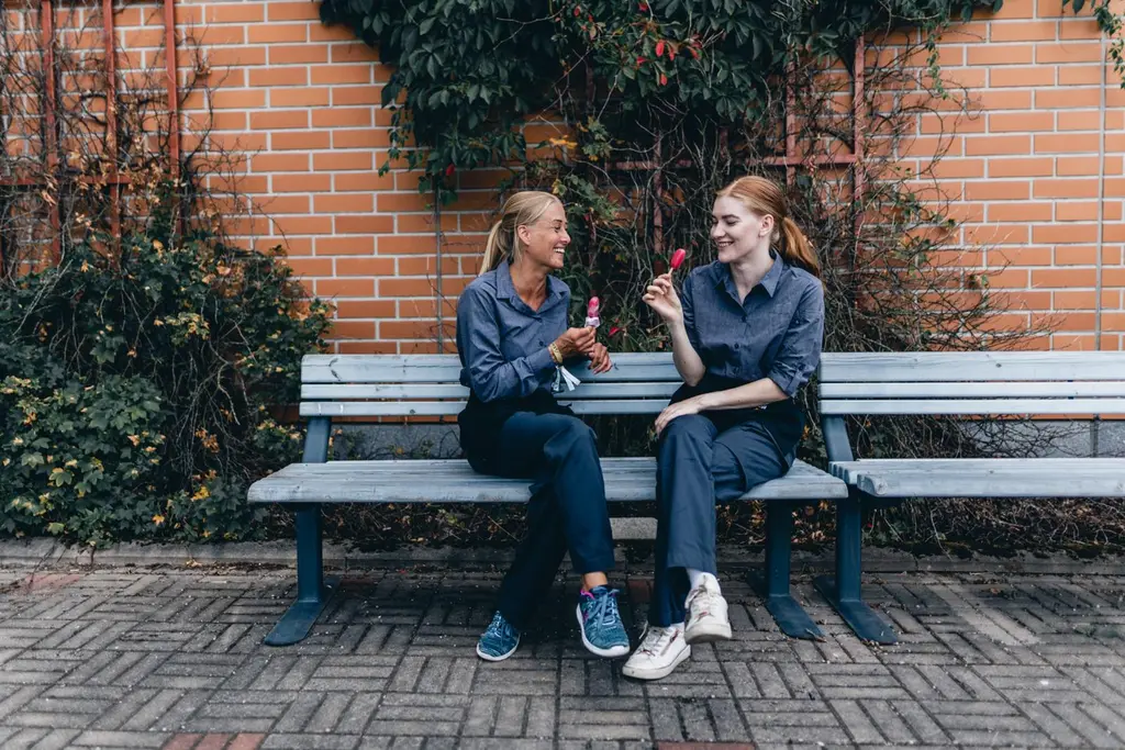 Kaksi ABC-työntekijää istuu liikenneaseman pihan penkillä. Heillä on käsissään mehujäät.