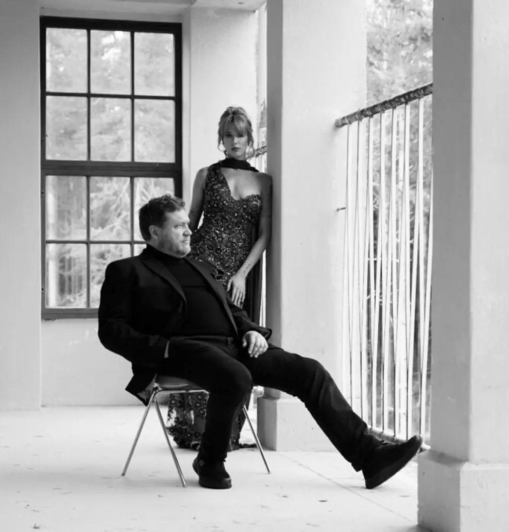 Mustavalkoinen kuva, jossa mies istuu tuolilla ja katsoo ulos ikkunasta. Hänen takanaan nainen iltapuvussa nojaa seinään ja katsoo kameraan