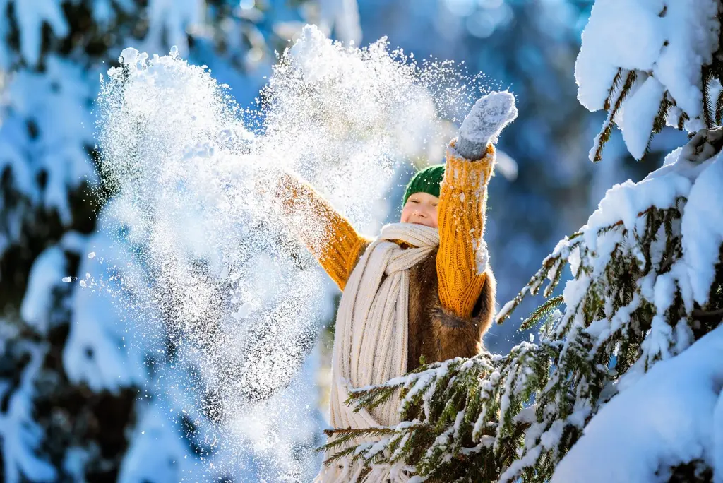Nuori keltaisessa villapaidassa heittää lunta ilmaan havupuiden keskellä.