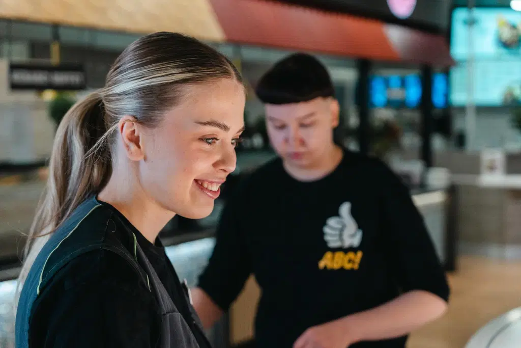 Nuori ABC-työntekijä hymyilee kuvassa etualalla. Työkaveri tekee taustalla työtehtävää.