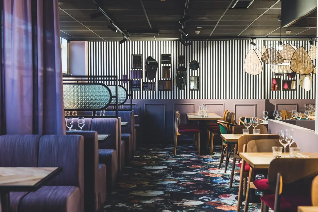 Ravintolan sisustus, jossa on värikäs kukkamatto, puupöytiä ja erivärisiä tuoleja. Mukava ja moderni tunnelma.