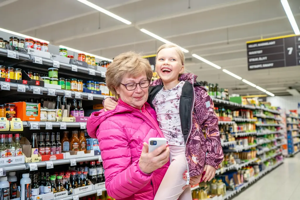Iäkkäämpi nainen katsoo puhelinta lapsi sylissään S-marketissa