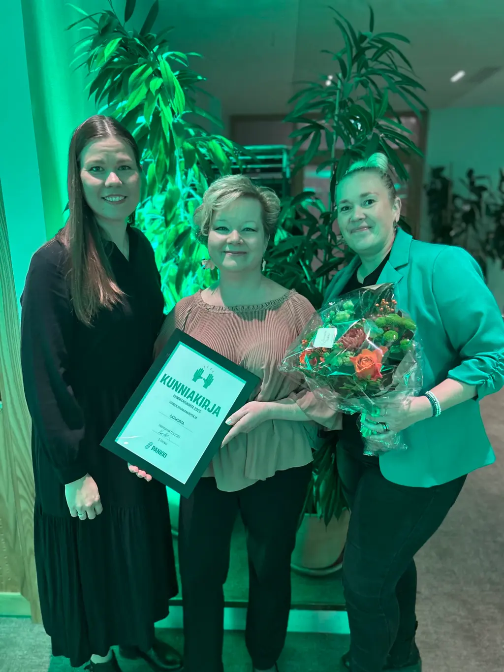 kolme naista esittelee kunniakirjaa ja kukkasia