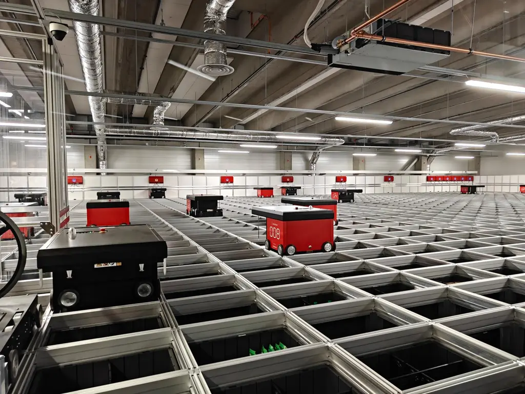 Automaattinen varastojärjestelmä, jossa punaisia kuljetusrobotteja liikkuu metallikourujen yläpuolella.