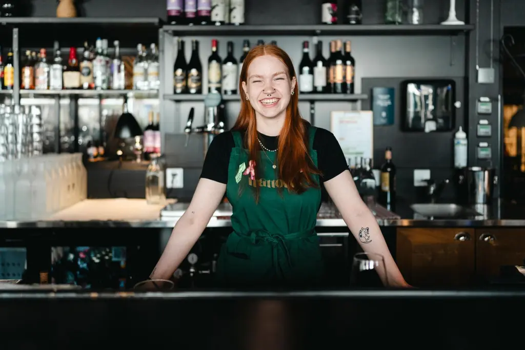 Seurahuone Bar & Bistron työntekijä seisoo tiskin takana essu päälään. Hän hymyilee leveästi.