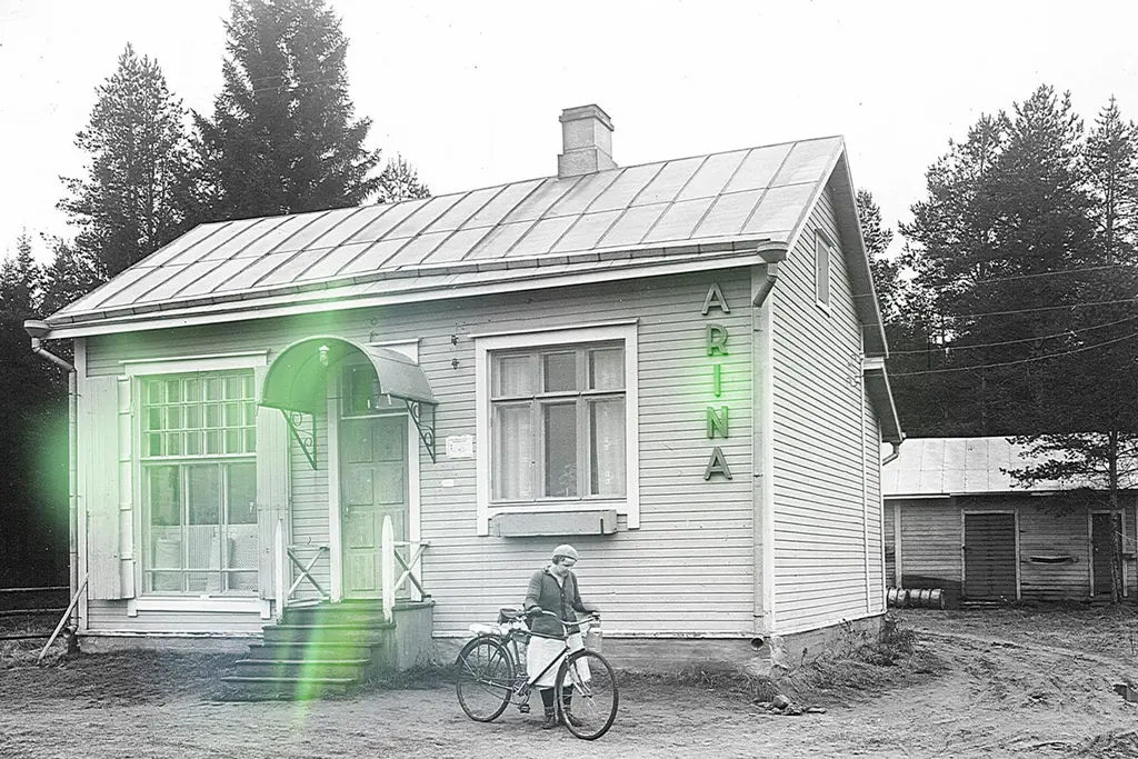 Puinen myymälärakennus, jonka seinässä teksti Arina. Kuvan etualalla tyttö pyörän kanssa