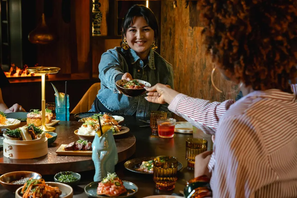 Nainen pitää kädessään ruoka-annosta, jota tarjoaa vastapäätä istuvalle ihmiselle.