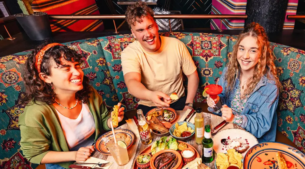 Kolme nuorta aikuista nauttivat iloisina texmex-ruoasta ravintolanpöydässä.