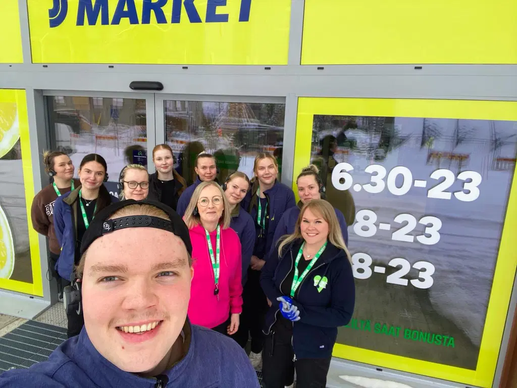 Joukko iloisia ihmisiä poseeraa kameralle S-marketin ulkopuolella.