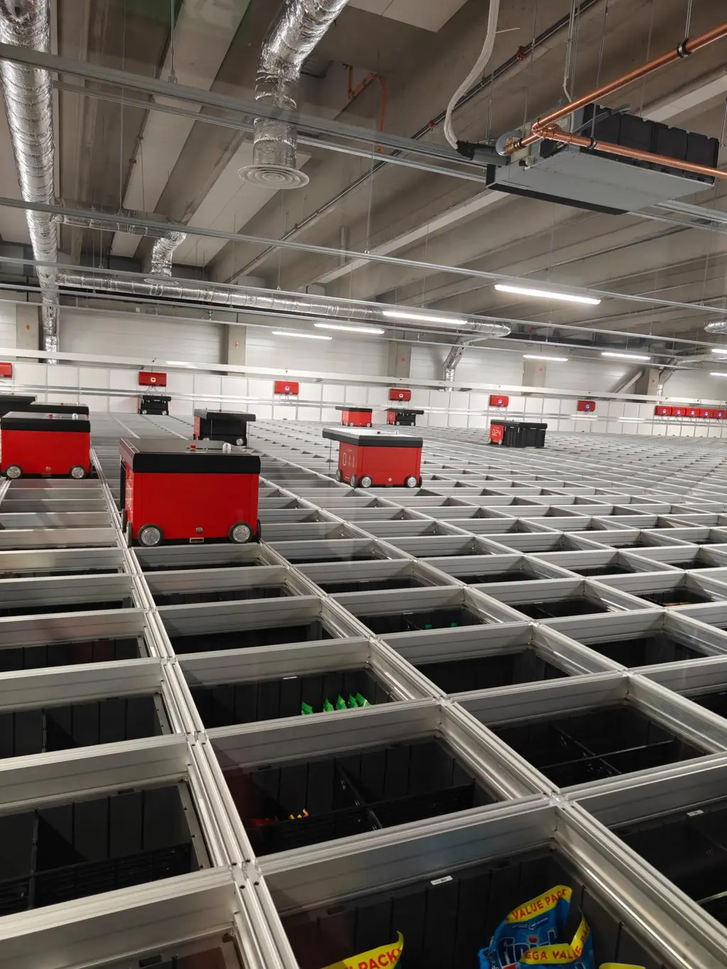 Automaattinen varastojärjestelmä, jossa punaisia kuljetusrobotteja liikkuu metallikourujen yläpuolella.