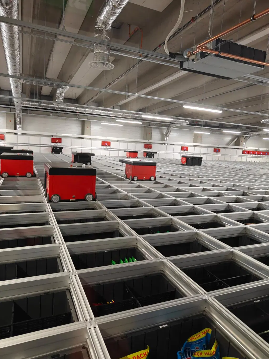 Automaattinen varastojärjestelmä, jossa punaisia kuljetusrobotteja liikkuu metallikourujen yläpuolella.