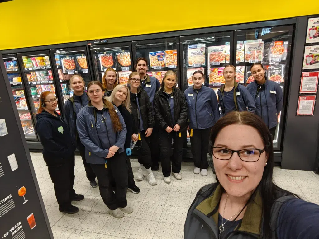 Joukko iloisia ihmisiä poseeraa kameralle S-marketin sisällä.