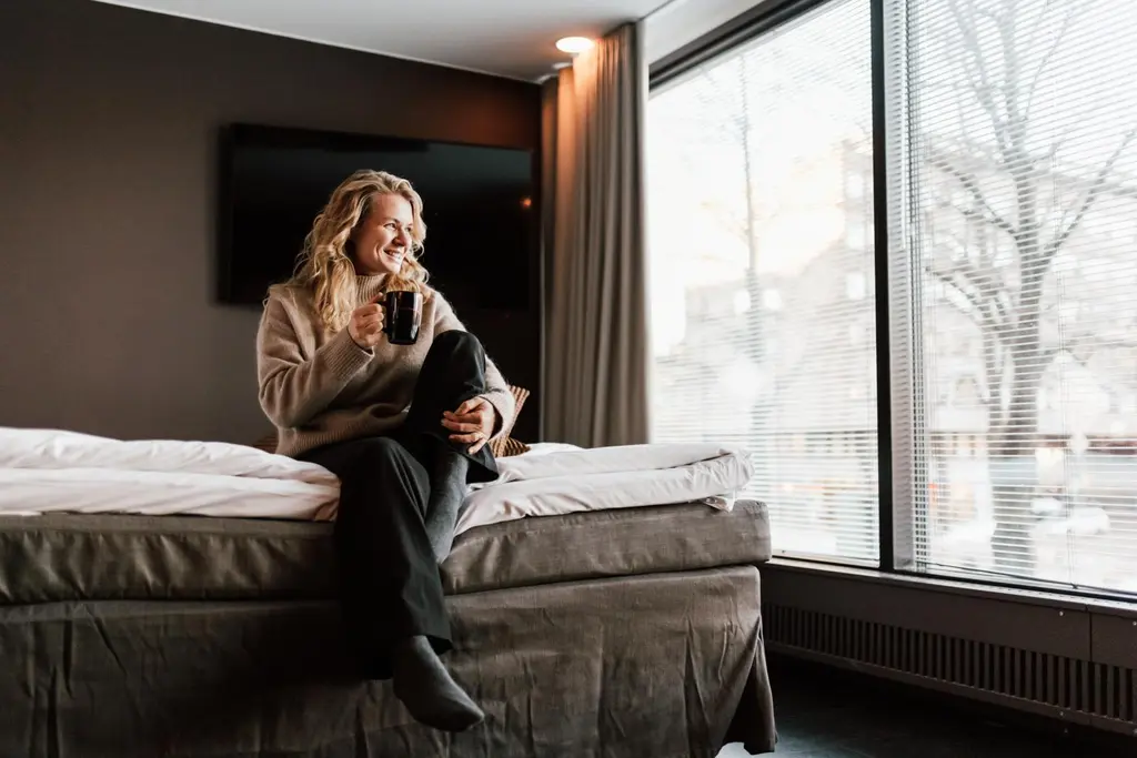 Nainen istuu kahvimukin kanssa hotellisängyn reunalla ja katsoo kaupunkimaisemaa ikkunasta.