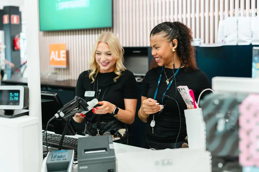 Kaksi Sokoksen työntekijää on kassapäätteellä. Toinen tarkistaa tuotteen hintaa.