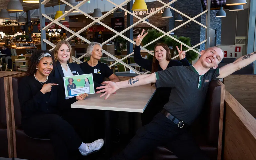 Viiden henkilön iloinen porukka istuu pöydän ääressä. Katseet on kameraan päin.