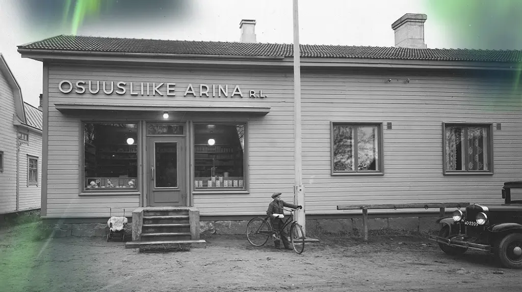 Vanha puinen myymälä, jonka seinässä kyltti Osuusliike Arina r.l. Myymälän edessä pieni poika pyörän kanssa