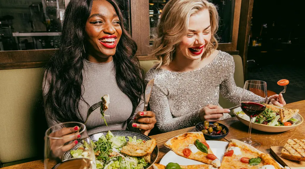 Kaksi iloista naista ravintolapöydässä pizzan ja salaatin ääressä.