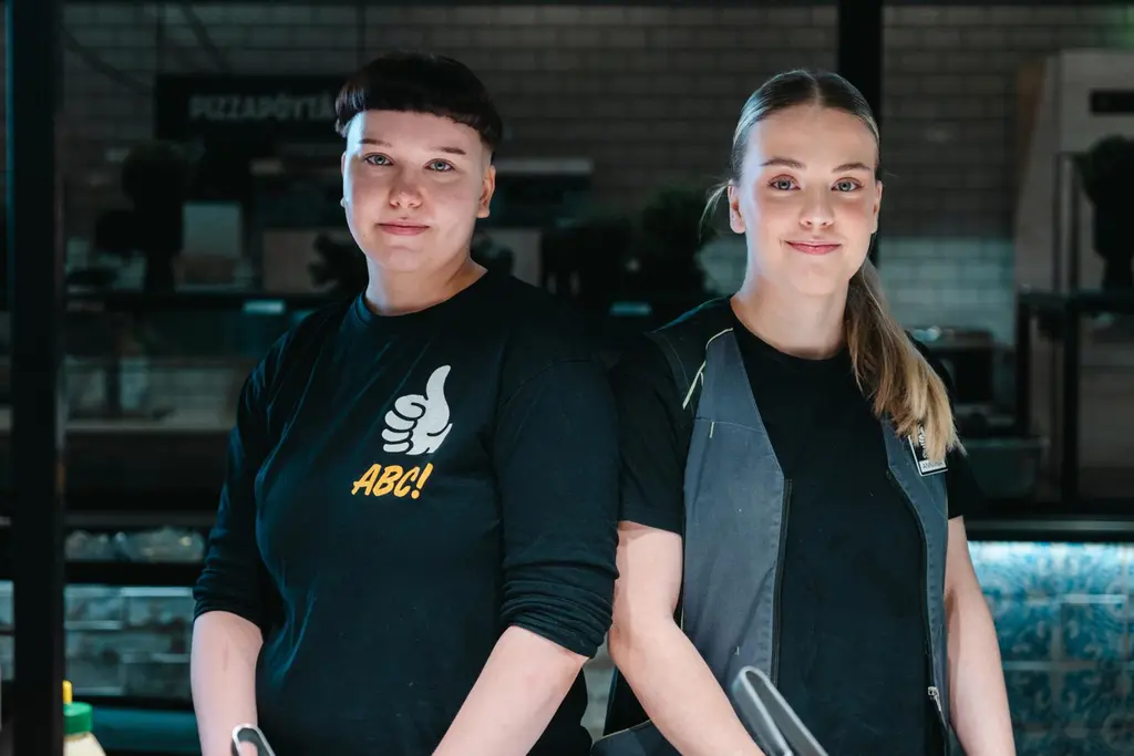 Kaksi nuorta ABC-työntekijää seisoo kasvot kameraan päin. Kumpikin hymyilee pienesti.