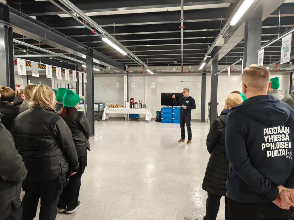 Ryhmä ihmisiä seisoo sisätiloissa kuuntelemassa puhujaa. Taustalla on pöytiä, joilla on tarjottavia ja juomia.