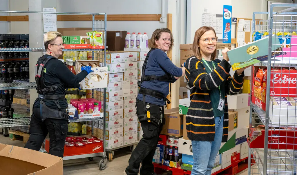 Kolme henkilöä purkaa kuormaa rullakoista ruokakaupan varastossa. Kahdella on käytössä eksoskeleton eli ulkoinen tukiranka.