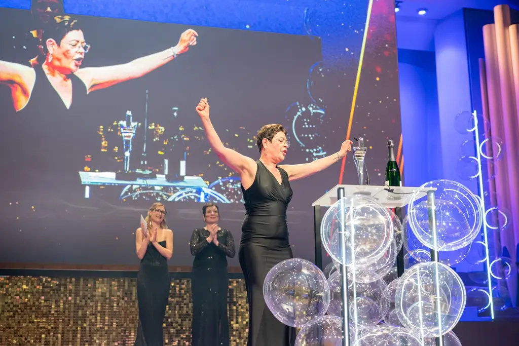 Nainen iltapuvussa nostaa käsiään ylös palkintolavalla, taustalla näyttö ja kaksi henkilöä taputtaa.