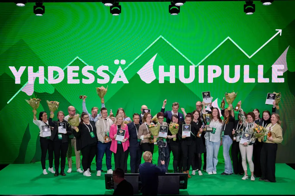 Joukko ihmisiä hurraa lavalla vihreässä tunnelmavalaistuksessa.