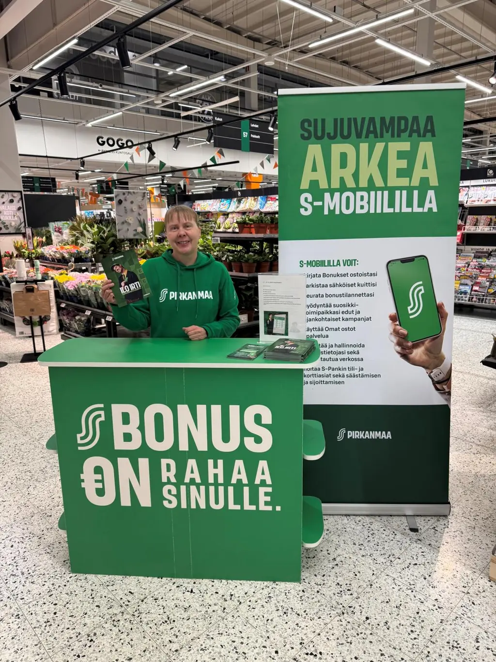 Kuvassa S-mobiilineuvoja hymyilemässä julisteen ja pöydän luona