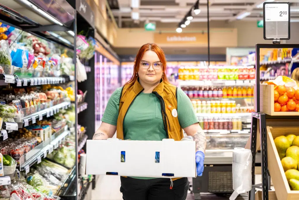 Salen työntekijä katsoo hymyillen kameraan pidellen käsissään vihanneslaatikkoa.