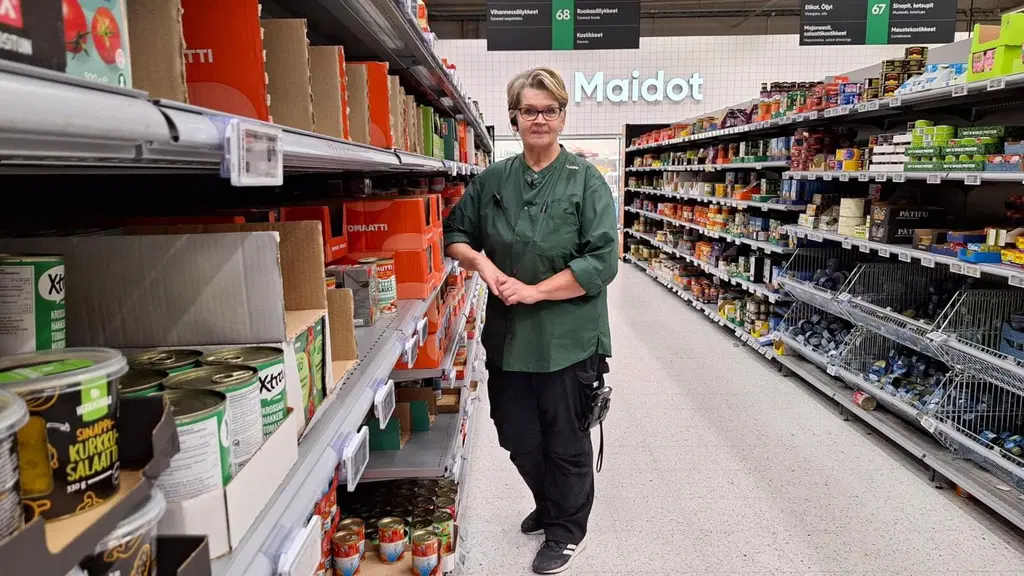 Yksi henkilö seisoo kaupan hyllyjen välissä. Taustalla on maitokaapit.
