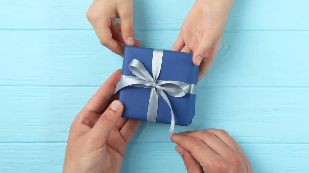 Pienet kädet ojentavat sinistä hopeisella lahjanarulla koristettu pakettia suuremmille käsille vaaleansinisen puisen pöydän päällä.
