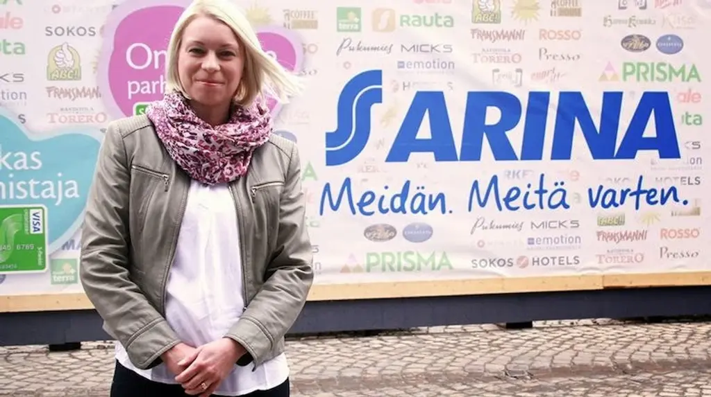 Vaalea nainen seisoo kyltin edessä ja katsoo hymyillen kameraan. Kyltissä teksti "S Arina Meidän. Meitä varten."