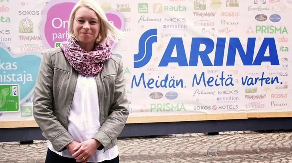 Vaalea nainen seisoo kyltin edessä ja katsoo hymyillen kameraan. Kyltissä teksti "S Arina Meidän. Meitä varten."