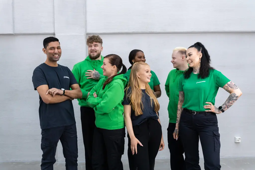 Ryhmä henkilöitä seisoo tiiviissä porukassa toisiaan katsoen ja hymyillen.