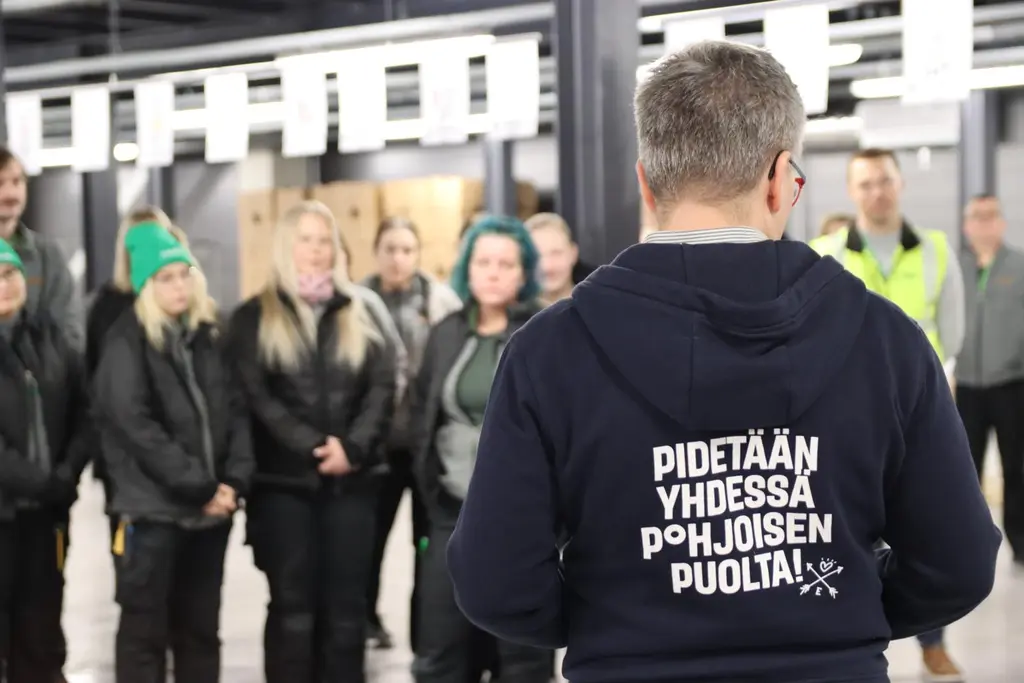 Mies takaapäin, hupparin selässä teksti "Pidetään yhdessä pohjoisen puolta". Taustalla ihmisiä kuuntelemassa miehen puhetta.