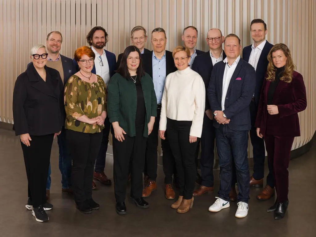 Kahdeksan miestä ja viisi naista seisoo puolikaaressa ryhmäkuvassa. He katsovat  kameraan ja hymyilevät. He ovat pukeutuneet business casual -tyyliin; osa käyttää bleisereitä, osa neuleita tai kuviollisia yläosia.