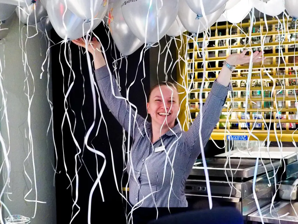 Iloinen työntekijä kohottaa kädet ilmaan ilmapalloryppään alla. 