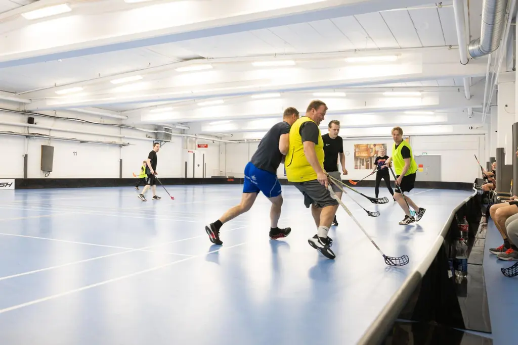 Joukko henkilöitä pelaa liikuntasalissa salibandya. Kuvan etualalla kamppaillaan pallosta.