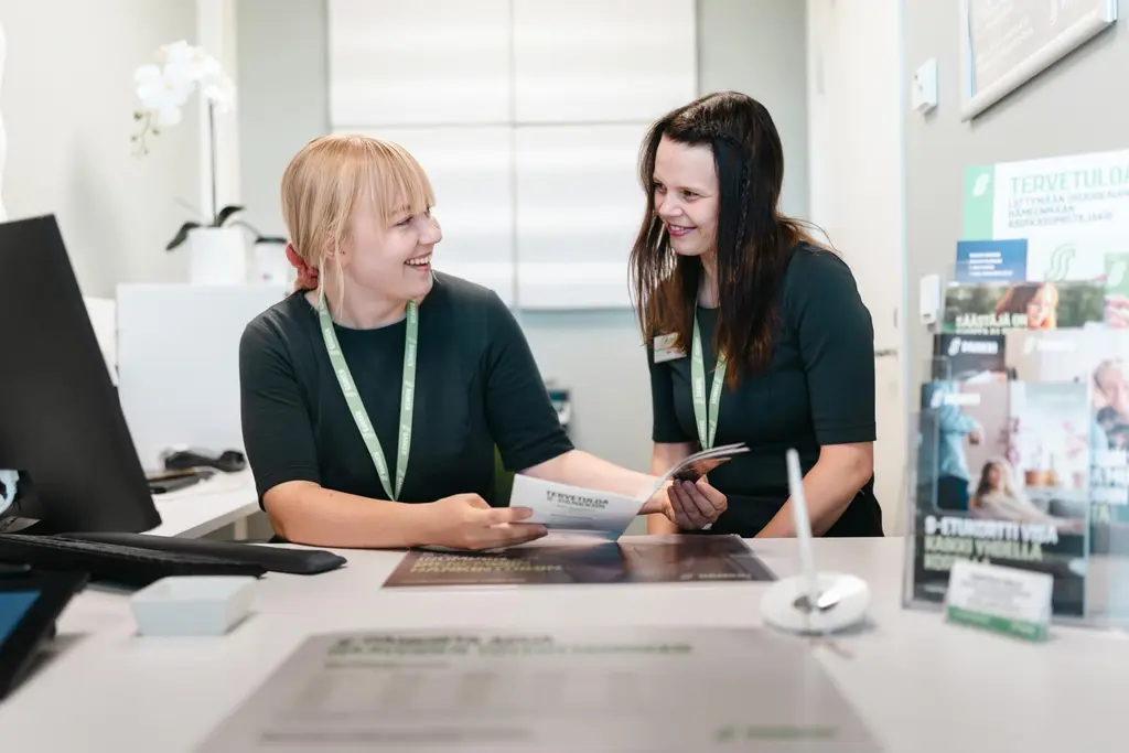 Kaksi S-Pankin työntekijää istuu pöydän takana ja katsoo toisiaan. Toisella on paperinen esite kädessä.