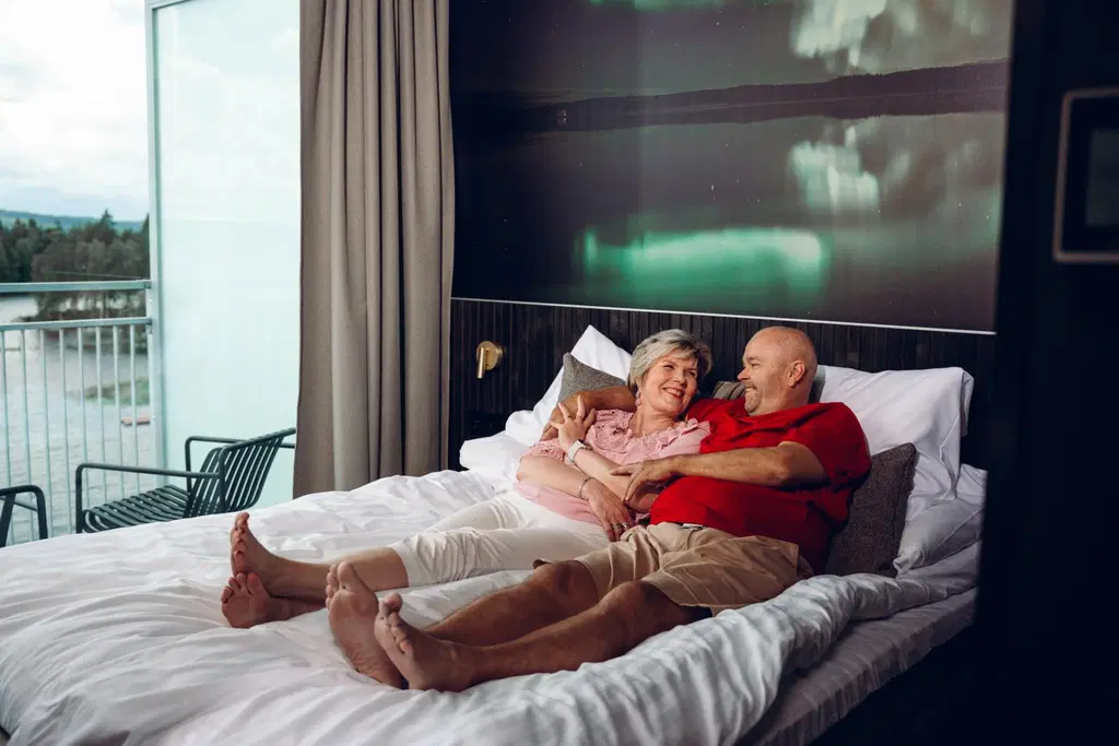 A couple is lying next to each other on a hotel bed, smiling.
