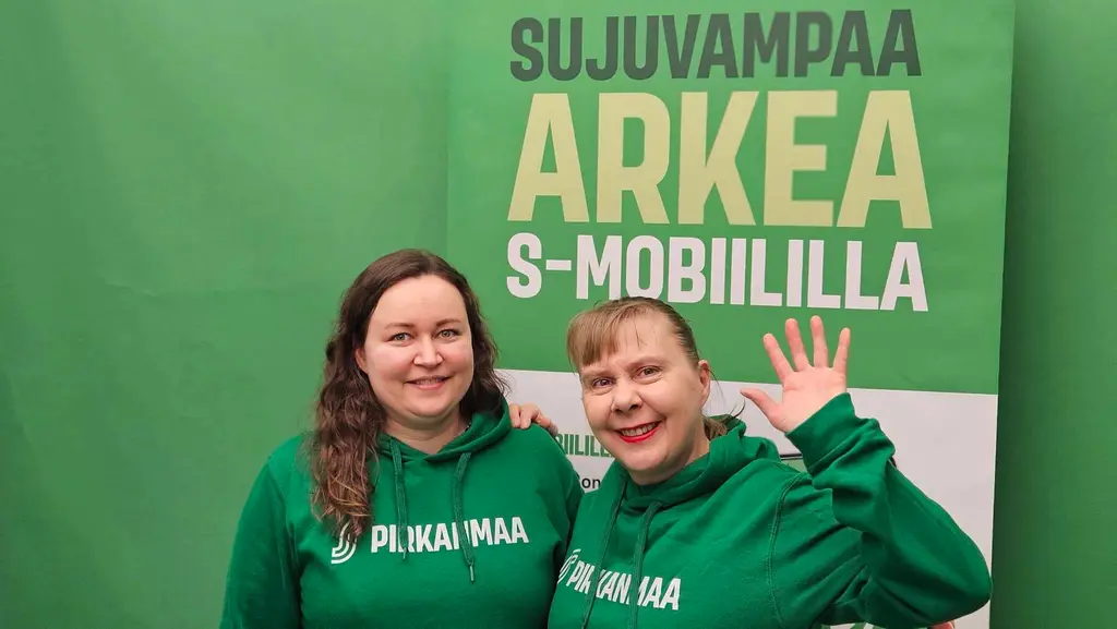 Kaksi S-mobiilineuvojaa tervehtii S-mobiili-banderollin edessä