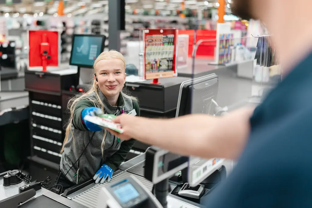 Kaupan kassamyyjä palvelee asiakasta, joka on kuvassa selkä kameraan päin.