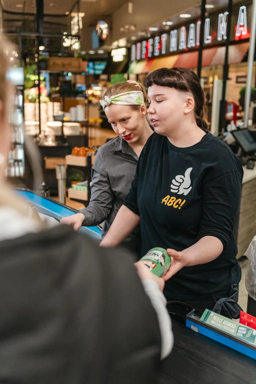 Kassalla on kaksi nuorta ABC-työntekijää myymässä tuotetta asiakkaalle. Perehdytystilanne.