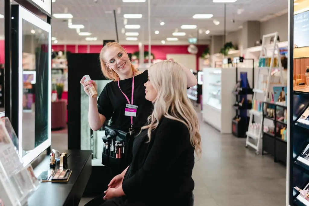 Sokoksen työntekijä meikkaa asiakasta peilin edessä. Kuva on otettu sivusta.