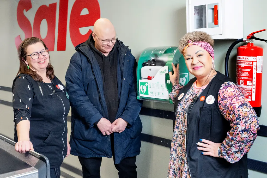 Kirsi, Markku ja Suvi defibrillaattorin vieressä. Defibrillaattorit kaikkiin Pirkanmaan Osuuskaupan toimipaikkoihin.