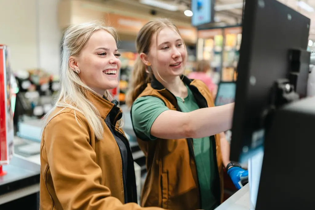 Kaksi nuorta työntekijää seisoo kassapäätteen edessä myymälässä. Toinen osoittaa jotain näytöltä.
